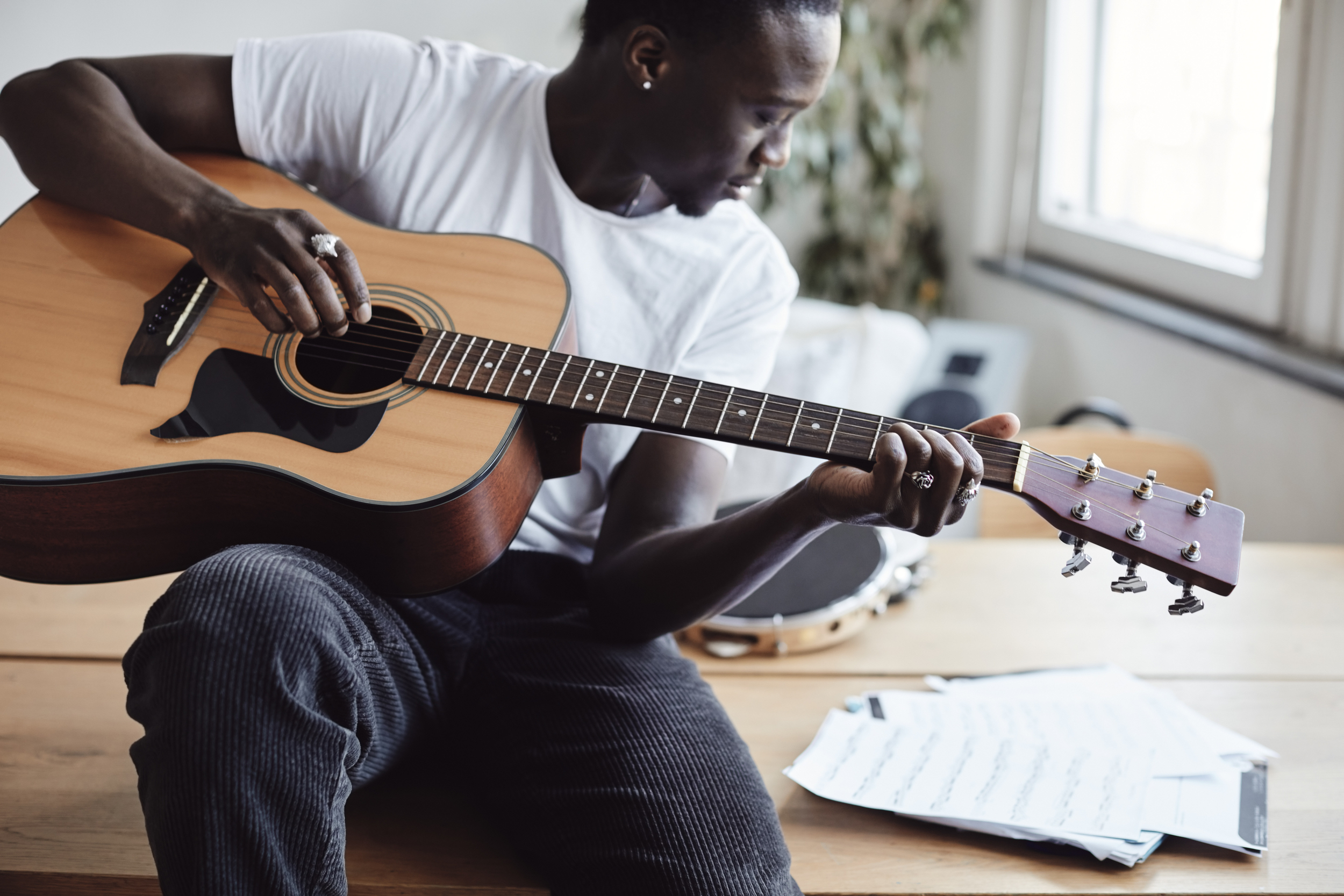 En kille i tonåren sitter på ett bord och spelar gitarr. Han har mörkt hår, vit tröja och svarta byxor. På bordet ligger det papper med noter. 
