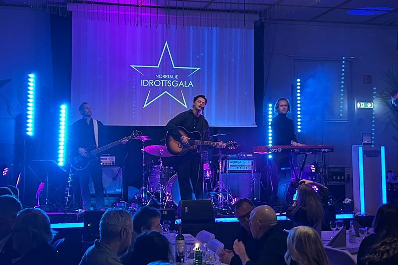 Tre killar står på en scen. Två spelar gitarr. En spelar keyboard