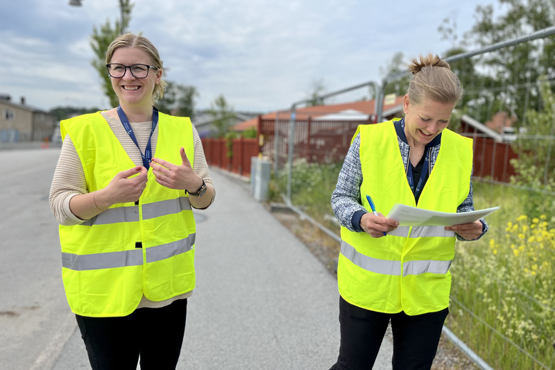 Tv&aring; kvinnor i reflexv&auml;star st&aring;r utomhus utanf&ouml;r ett byggstaket och ler.