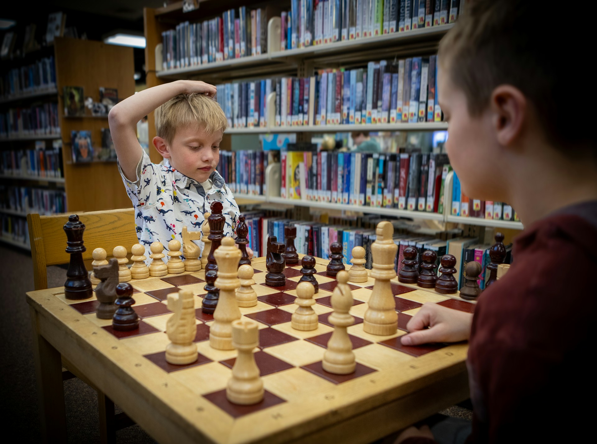 Två barn spelar schack vid ett stort träschackbräde i ett bibliotek.