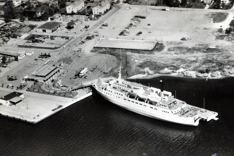 Historisk svartvit bild &ouml;ver Norrt&auml;lje hamns f&auml;rjel&auml;ger. Bilden &auml;r tagen uppifr&aring;n och det ligger en stor f&auml;rja i vattnet. P&aring; land st&aring;r bilar parkerade.