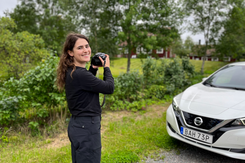 Isabelle Sahba, en kvinna i trettioårsåldern med mörka kläder och mörkt, lockigt hår står med en kamera framför en vit bil, tittar in i kameran och ler.