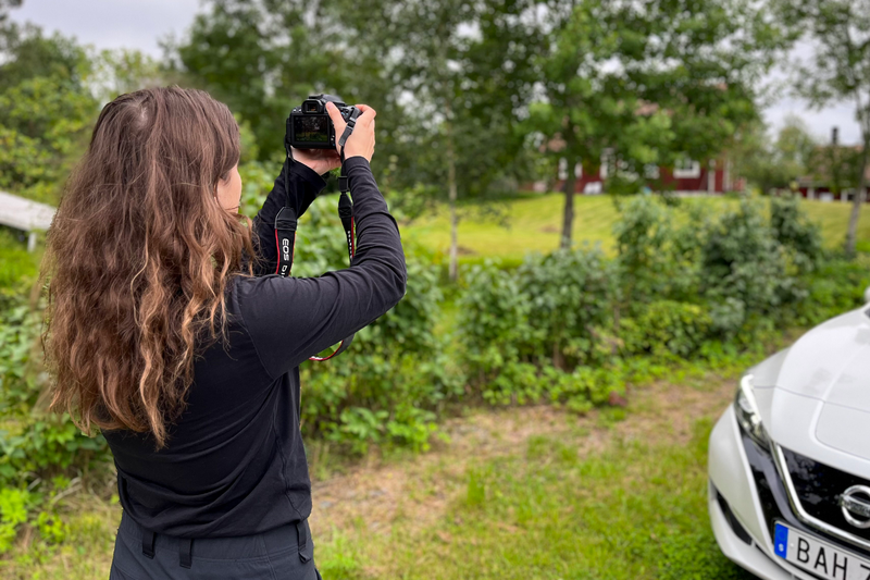 Isabelle Sahba, en kvinna i trettio&aring;rs&aring;ldern med m&ouml;rka kl&auml;der och m&ouml;rkt, lockigt h&aring;r st&aring;r med en kamera framf&ouml;r en vit bil och fotograferar ett r&ouml;tt hus.