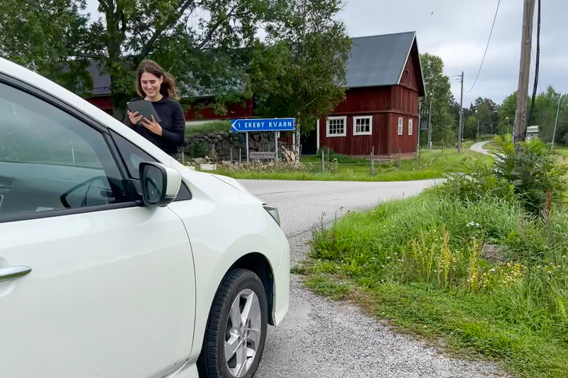 Isabelle Sahba, en kvinna i trettio&aring;rs&aring;ldern med m&ouml;rka kl&auml;der och m&ouml;rkt, l&aring;ngt, lockigt h&aring;r st&aring;r med en kamera framf&ouml;r en vit bil.