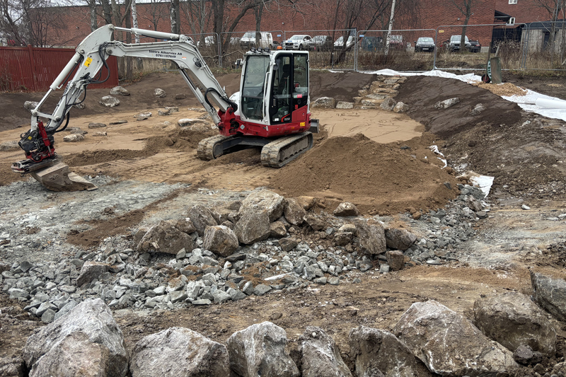 Gr&auml;vmaskin som jobbar i en grop med sand, stora stenar runtomkring och en stentrappa ner.