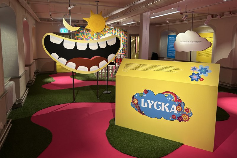 Bilden visar entren av utst&auml;llning Lycka. En mun som skrattar, en sol och en m&aring;ne. Golvet &auml;r rosa och gr&ouml;nt.