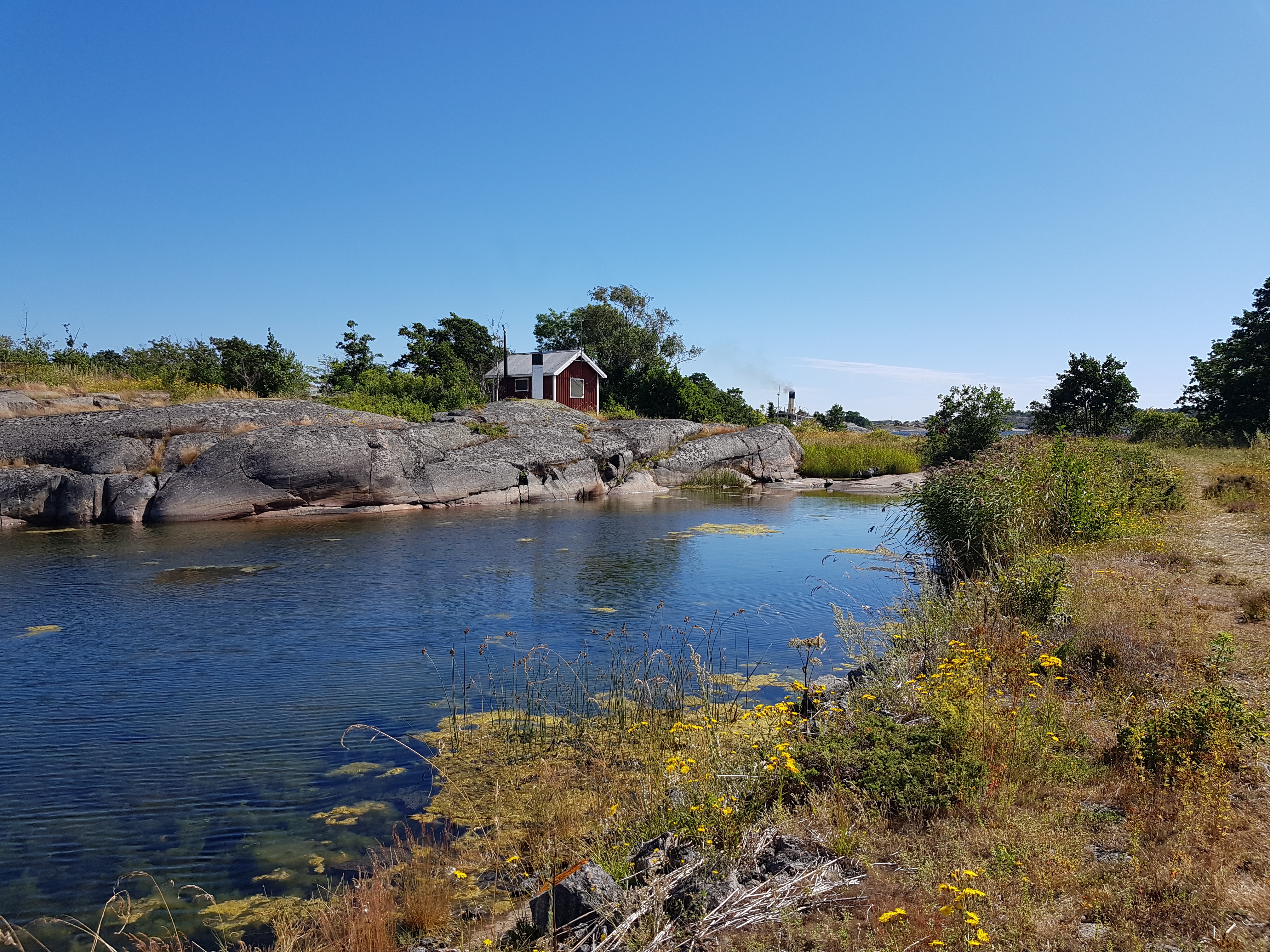 På bilden syns ön Båtskären. I bilden vatten, klippor med en röd fiskebod, en blå himmel, grönska och längst bak i bilden syns röken från S/S Blidösund.