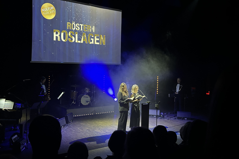 Tv&aring; kvinnor st&aring;r p&aring; en scen och l&auml;ser ur en bok