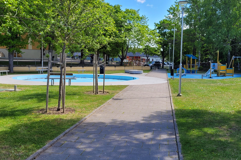 Bilden visar Plaskdammsparken i Hallstavik. P&aring; bilden ser man planteringar med perenner och parkb&auml;nkar d&auml;r man kan vila sig i skuggan av ett tr&auml;d. I parken finns &auml;ven en plaskdamm och en lekpark.