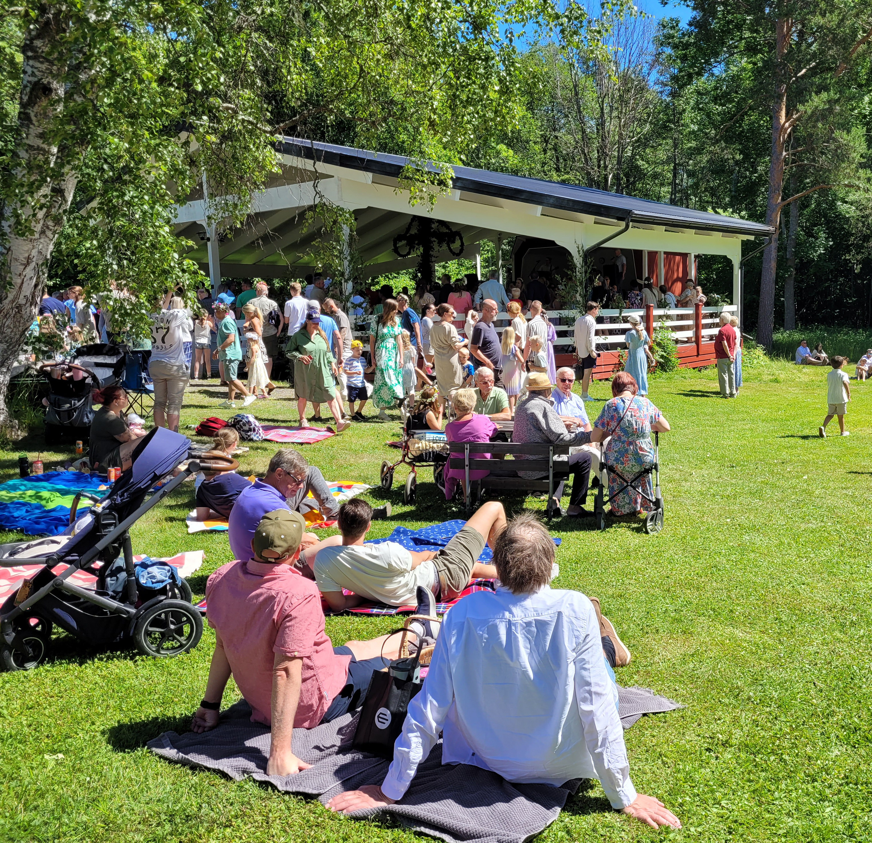 Dansbana med midsommarfirande och människor i gröngräset framför.
