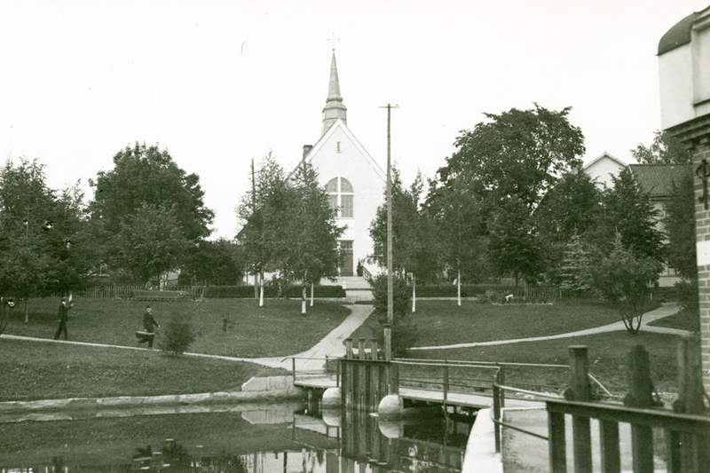 Historisk bild &ouml;ver Norrt&auml;lje &aring;n. I f&ouml;rgrunden syns en liten bro &ouml;ver &aring;n, i bakgrunden tornar en vit kyrkoliknande byggnad upp sig.