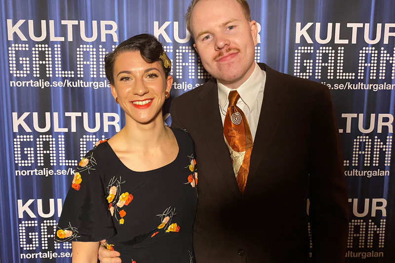 Dansarna Bianca och Nils visas på bilden. Bianca har en svart klänning med blommor och Nils har en svart kostym och guld/orange slips.