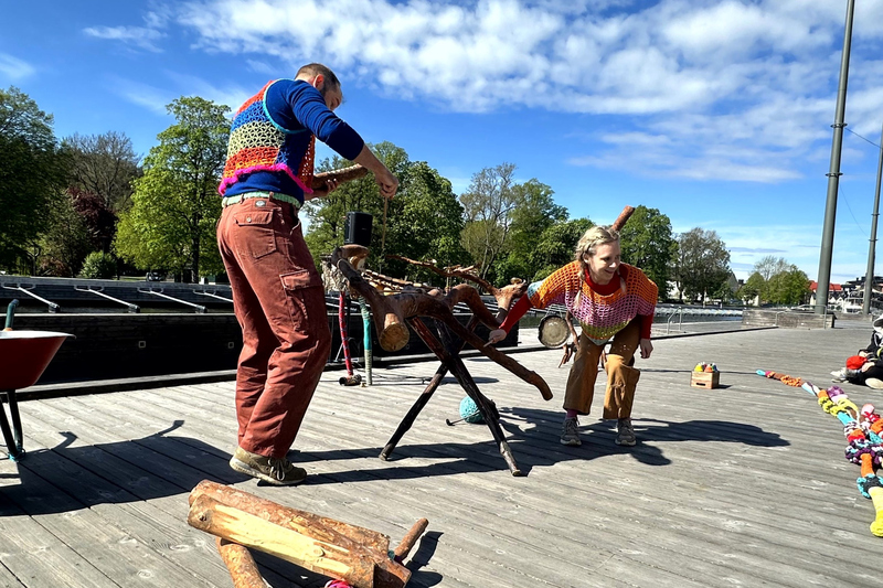En man och en kvinna i f&auml;rgglada virkade kl&auml;der dansar och trummar p&aring; en tr&auml;dgren