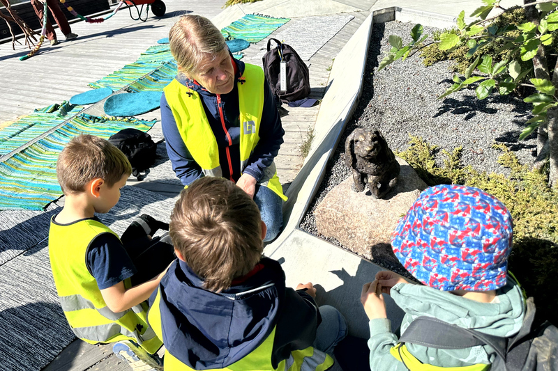 tre barn och en pedagog i reflexv&auml;star sitter runt en kanin i brons och pratar